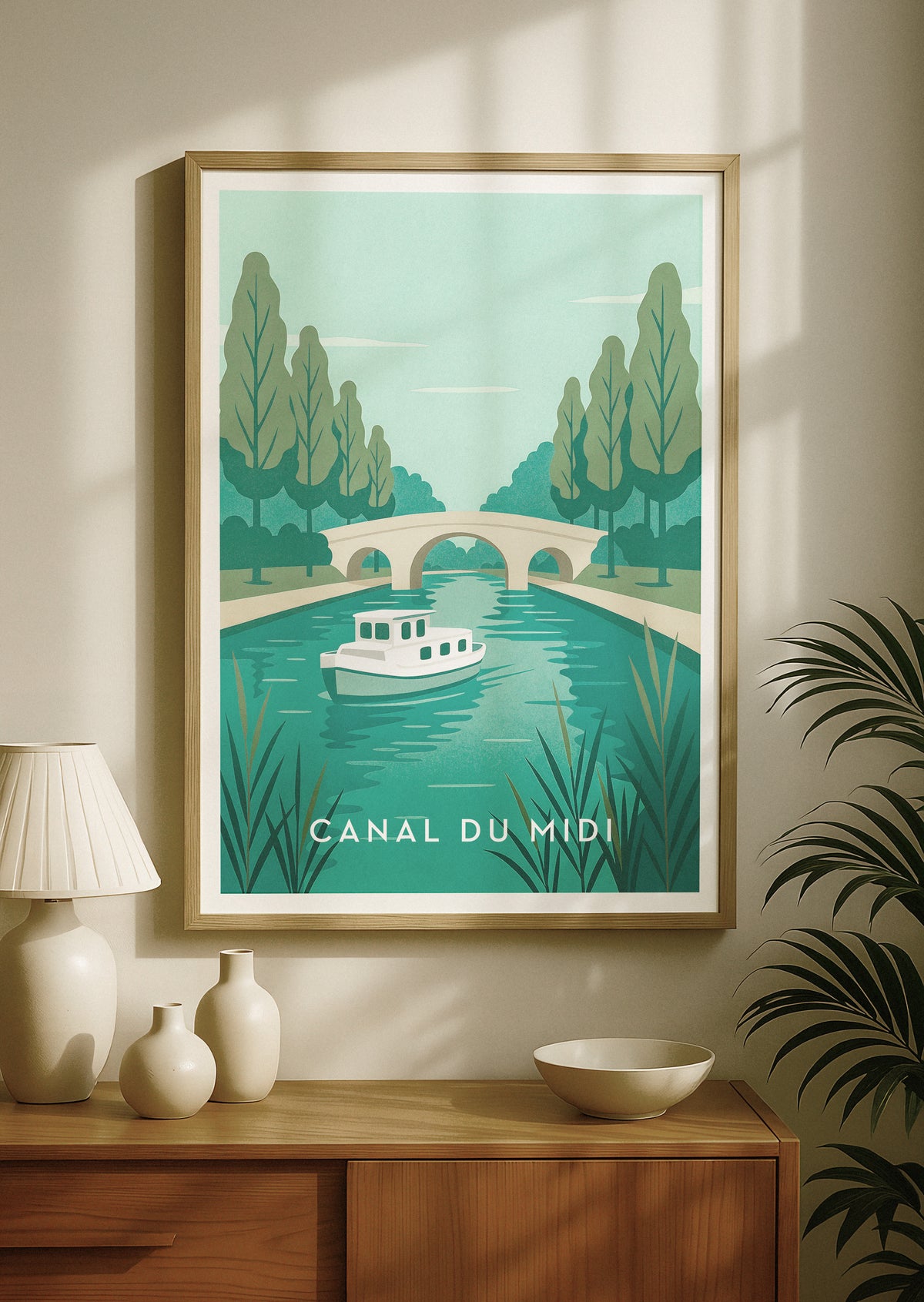 Kehystetty, matkailujulistetyylinen juliste Canal du Midistä Ranskassa. Kuvassa on retrolaiva turkoosilla vedellä, puita ja kivisilta. Juliste on esillä beigenvärisellä seinällä puisen konsolipöydän yläpuolella, pöydällä koristeellisia maljakoita ja lamppu.